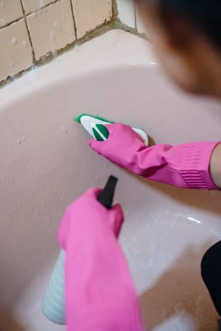 gallery-03 Personne anonyme portant des gants en caoutchouc jaunes et utilisant une simple éponge pour nettoyer un miroir dans une salle de bain
